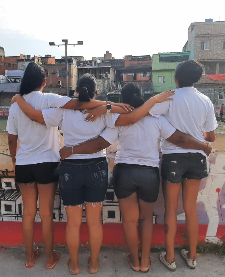 Quatro mulheres integrantes do Grupo Mães de Manguinhos aparecem de costas, abraçadas e olhado para a favela à sua frente